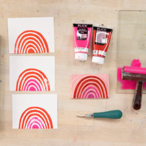flat lay on a wooden table of supplies for carving stamps, including 2 brayers, block printing ink in pink and red, a tray full of hand-carved rubber stamps, and colorful rainbow prints