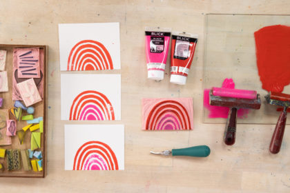 flat lay on a wooden table of supplies for carving stamps, including 2 brayers, block printing ink in pink and red, a tray full of hand-carved rubber stamps, and colorful rainbow prints