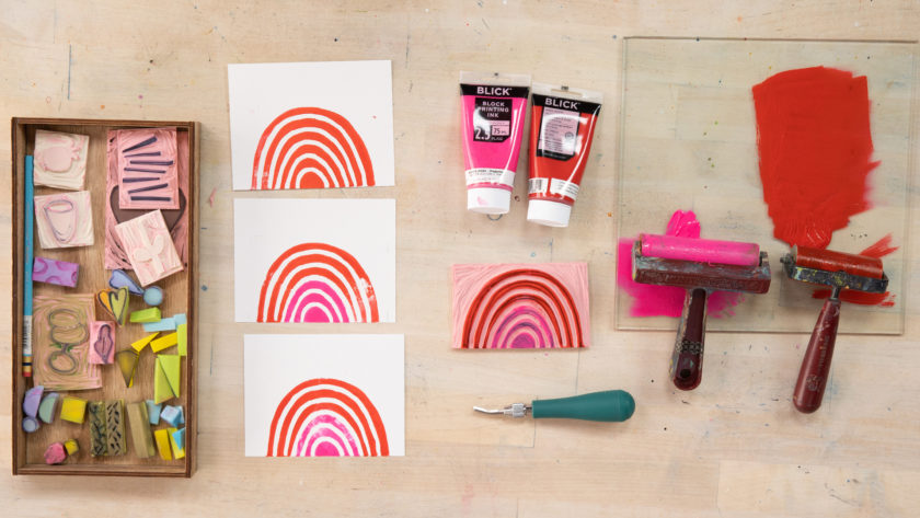 flat lay on a wooden table of supplies for carving stamps, including 2 brayers, block printing ink in pink and red, a tray full of hand-carved rubber stamps, and colorful rainbow prints