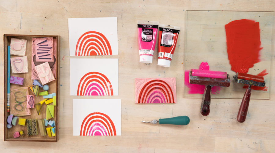 flat lay on a wooden table of supplies for carving stamps, including 2 brayers, block printing ink in pink and red, a tray full of hand-carved rubber stamps, and colorful rainbow prints
