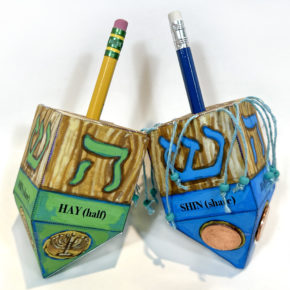 two handmade dreidels made of paper on a white background, one green, one blue, with pencils as handles