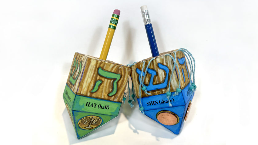 two handmade dreidels made of paper on a white background, one green, one blue, with pencils as handles