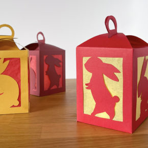 3 red and gold handmade paper lanterns with cut-outs of rabbits on a wooden tabletop