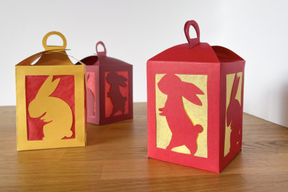 3 red and gold handmade paper lanterns with cut-outs of rabbits on a wooden tabletop