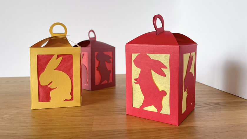 3 red and gold handmade paper lanterns with cut-outs of rabbits on a wooden tabletop