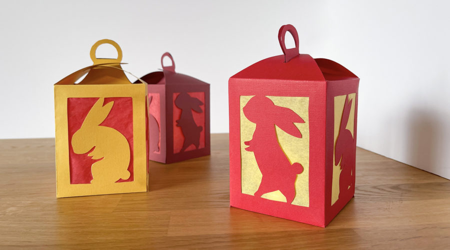 3 red and gold handmade paper lanterns with cut-outs of rabbits on a wooden tabletop