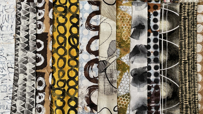 an array of folded fabrics lined up on a table, in black, white, and a some yellow, and bold, hand-painted strokes and patterns, designed by artist e bond