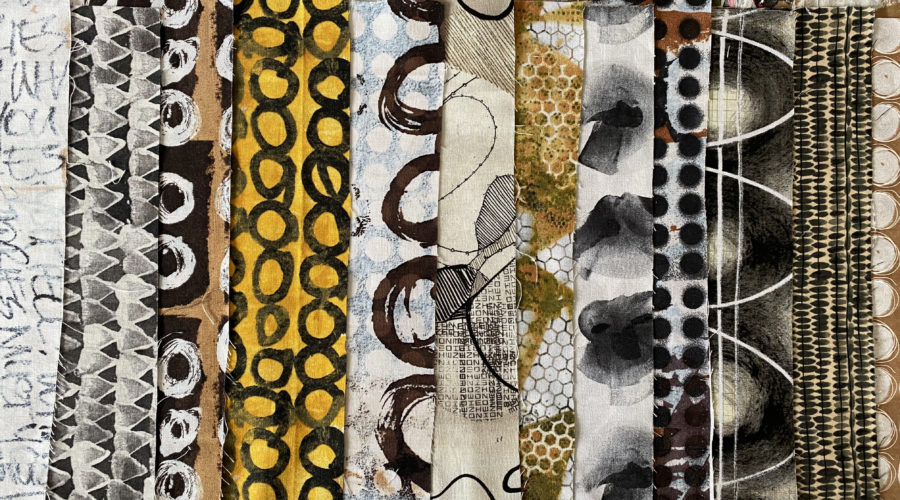 an array of folded fabrics lined up on a table, in black, white, and a some yellow, and bold, hand-painted strokes and patterns, designed by artist e bond