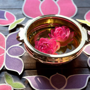 close-up photo of gold bowl with 2 pink flowers floating inside, surrounded by rangoli-inspired sand art in teh shapes of lotus flowers for Holi