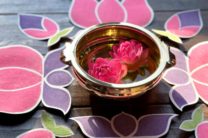 close-up photo of gold bowl with 2 pink flowers floating inside, surrounded by rangoli-inspired sand art in teh shapes of lotus flowers for Holi