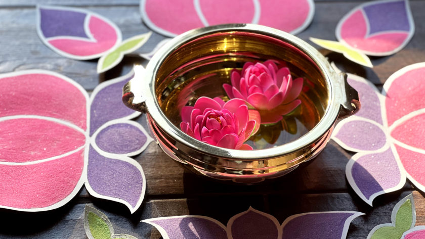 close-up photo of gold bowl with 2 pink flowers floating inside, surrounded by rangoli-inspired sand art in teh shapes of lotus flowers for Holi