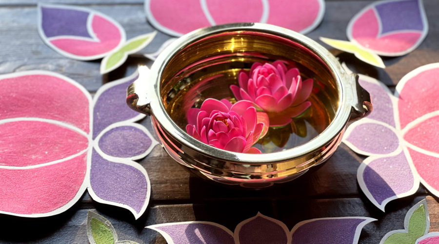 close-up photo of gold bowl with 2 pink flowers floating inside, surrounded by rangoli-inspired sand art in teh shapes of lotus flowers for Holi