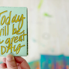 closeup image of a hand holding a small light green booklet that reads in yellow handwritten font "today will be a great day"