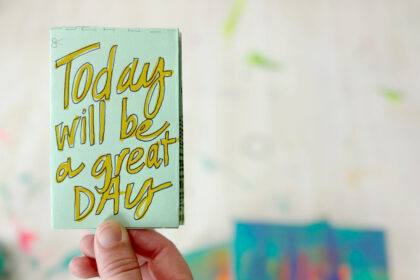 closeup image of a hand holding a small light green booklet that reads in yellow handwritten font "today will be a great day"
