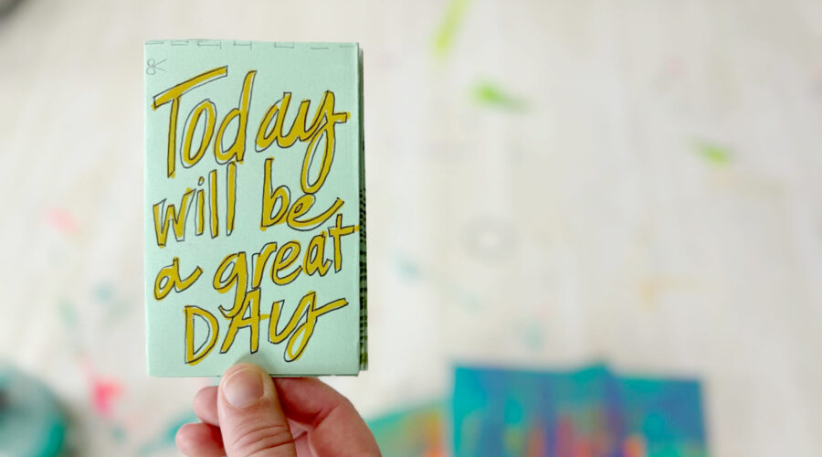 closeup image of a hand holding a small light green booklet that reads in yellow handwritten font "today will be a great day"