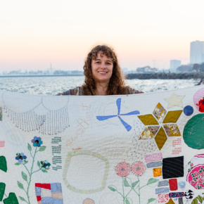 image of a woman holding up her hand stitched quilt by a waterfront