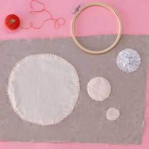 image of piece of grey fabric with a large white circle applique and smaller circle appliques, styled with red embroidery thread and an embroidery hoop all on a pink background