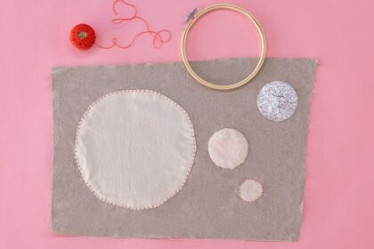 image of piece of grey fabric with a large white circle applique and smaller circle appliques, styled with red embroidery thread and an embroidery hoop all on a pink background