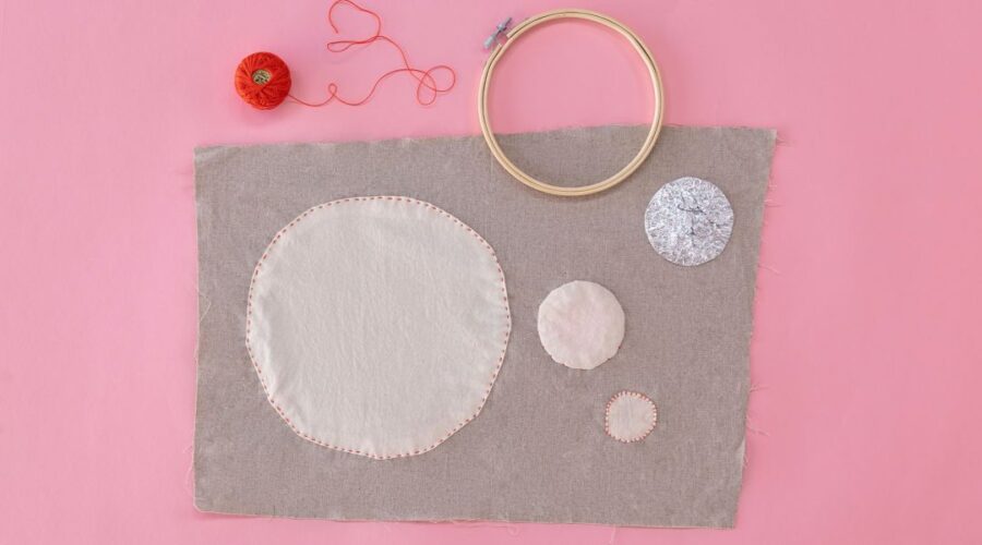 image of piece of grey fabric with a large white circle applique and smaller circle appliques, styled with red embroidery thread and an embroidery hoop all on a pink background