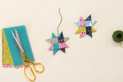 image of 2 woven fabric ornament stars on a cream colored background, styled with a ball of green string, a pair of scissors, and a colorful pile of fabric, to introduce handmade holiday gift ideas