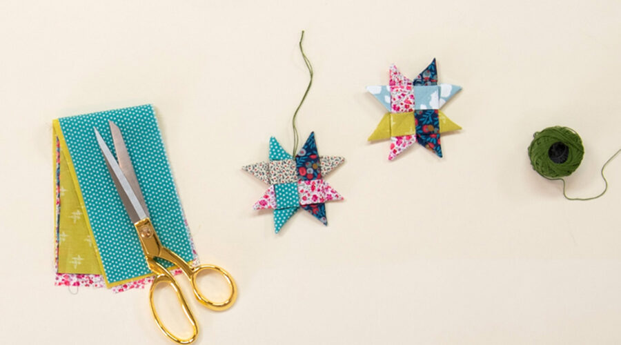 image of 2 woven fabric ornament stars on a cream colored background, styled with a ball of green string, a pair of scissors, and a colorful pile of fabric, to introduce handmade holiday gift ideas