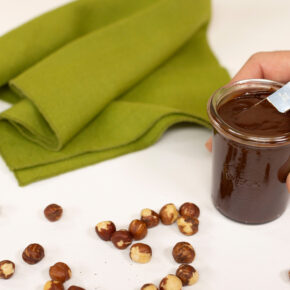 image of a hand holding a small jar of homemade chocolate hazelnut spread with some scattered hazelnuts on a table beside a green napkin