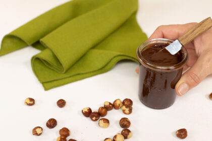 image of a hand holding a small jar of homemade chocolate hazelnut spread with some scattered hazelnuts on a table beside a green napkin