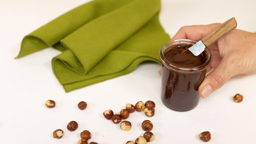 image of a hand holding a small jar of homemade chocolate hazelnut spread with some scattered hazelnuts on a table beside a green napkin