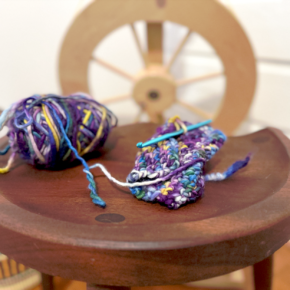 image of small knitting project and ball of yarn in blue and purple hues on a wood table with a wood spinning wheel in the background
