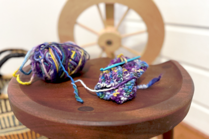image of small knitting project and ball of yarn in blue and purple hues on a wood table with a wood spinning wheel in the background