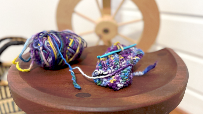 image of small knitting project and ball of yarn in blue and purple hues on a wood table with a wood spinning wheel in the background