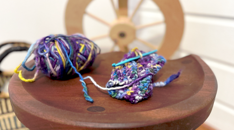 image of small knitting project and ball of yarn in blue and purple hues on a wood table with a wood spinning wheel in the background